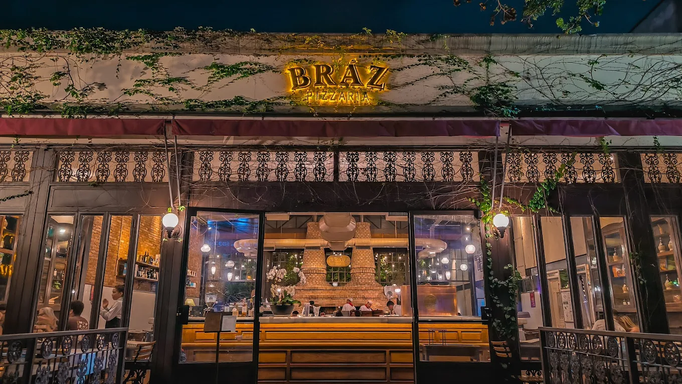 Fachada noturna da Bráz Pizzaria com iluminação amarela, parede com hera e design clássico