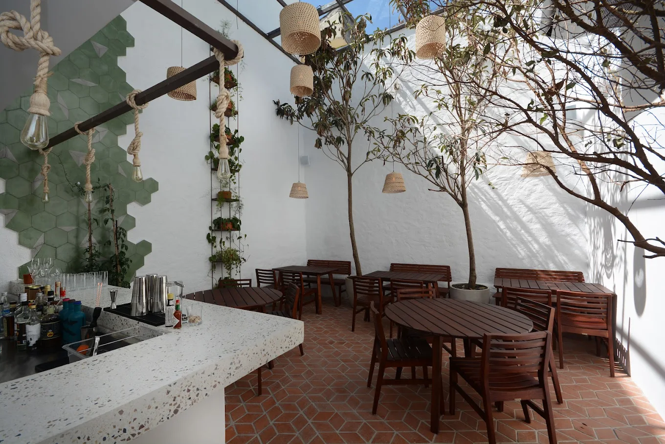 Área externa da Casa Rios no Tatuapé com mesas de madeira, árvores internas, luminárias de palha e bar com acabamento em terrazzo.