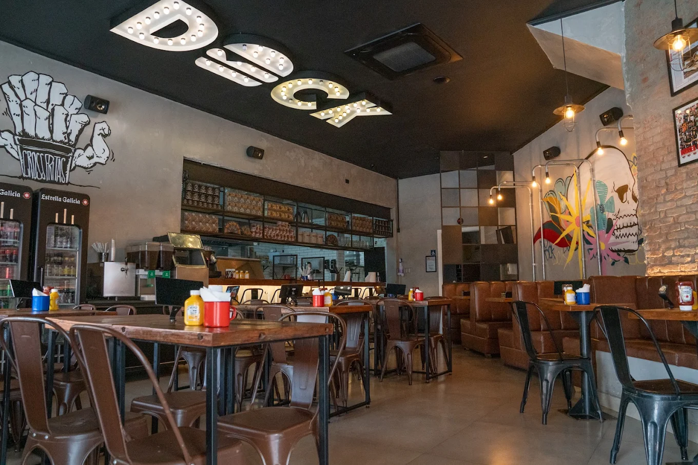 Interior do DCK Burguer com mesas industriais, parede de tijolos, arte urbana e letreiro iluminado no teto.