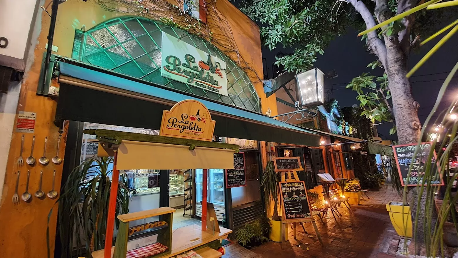 Fachada do restaurante La Pergoletta no Tatuapé, iluminada à noite, com decoração charmosa e cardápios na calçada.