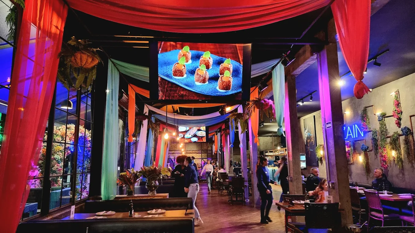 Interior colorido do restaurante Zain Tatuapé com decoração vibrante de tecidos vermelhos e turquesas, iluminação roxa e tela exibindo pratos gourmet.